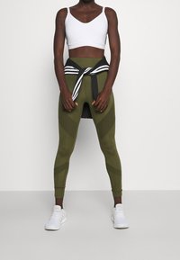 Witte sportbeha en olijfgroene leggings met geometrisch ontwerp. Zwart-witte gestreepte jas geknoopt om de taille. Witte sportschoenen.
