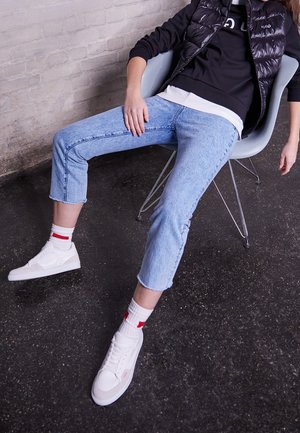 Person iført lyseblå cropped jeans, hvide sneakers med hvide sokker, en sort sweatshirt og en sort dunvest, der sidder afslappet på en stol.