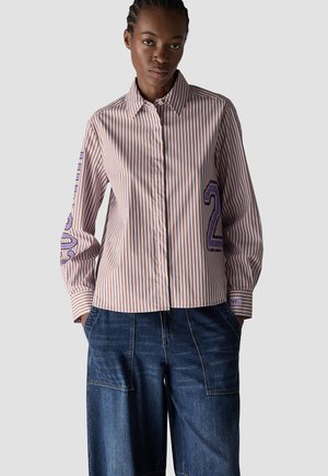 Woman wearing a brown and white striped button-up shirt with purple number patches on sleeves and dark blue loose jeans, against a plain background.