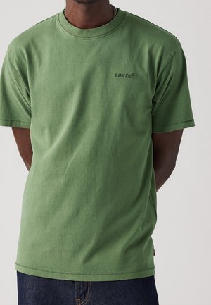 Mand iført en ensfarvet olivenfarvet Levi's t-shirt med korte ærmer, parret med mørkeblå jeans mod en ensfarvet baggrund.