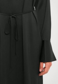 Rochie lungă de culoare neagră, cu mâneci evazate și o curea subțire legată în talie, având o mână odihnindu-se lângă articolul vestimentar.