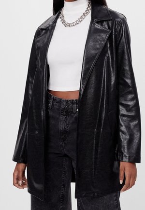 Frau trägt schwarzen Kunstledermantel über weißem, gekürztem Rollkragenpullover und schwarzen, hochgeschnittenen Jeans mit silberner Kettenkette.