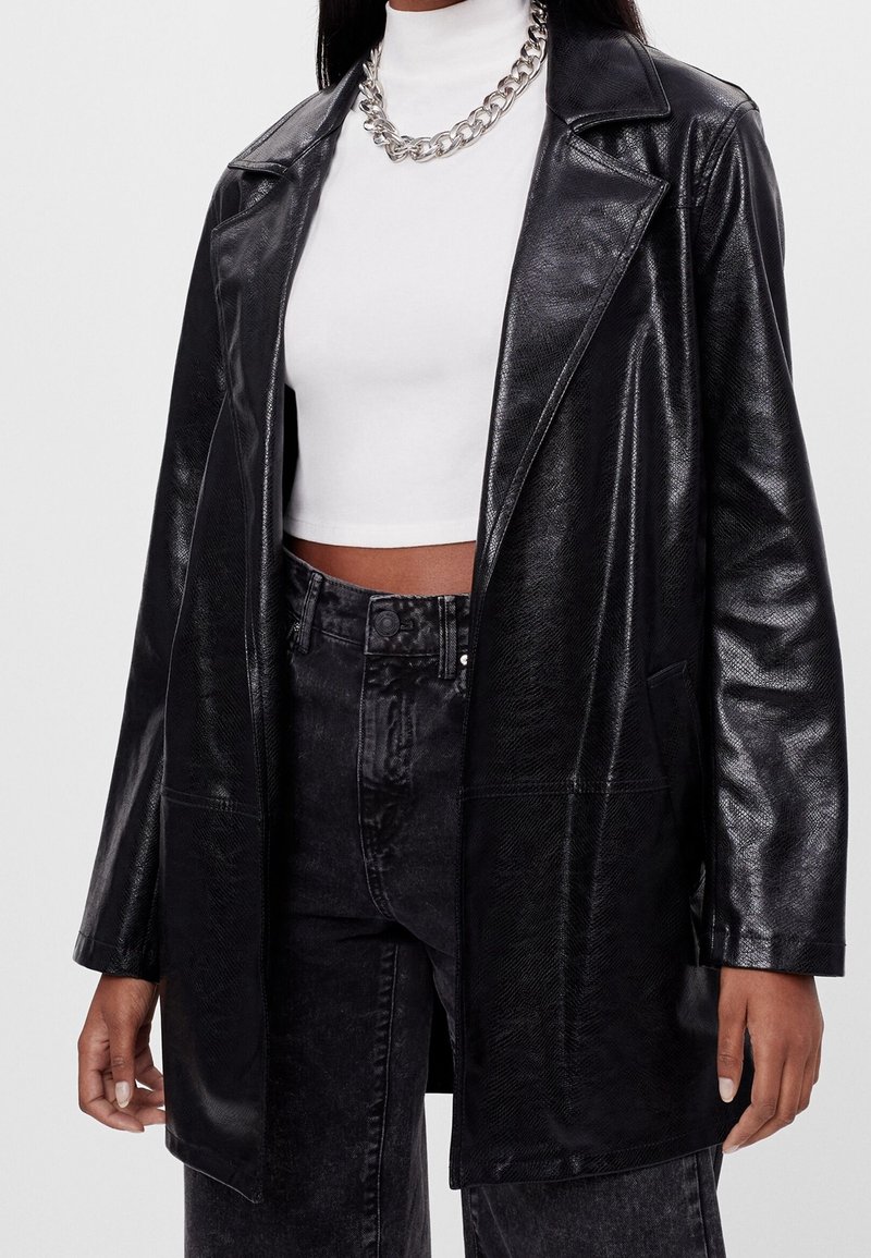 Femme portant un manteau noir en simili cuir sur un col roulé court blanc et un jean noir taille haute avec un collier chaîne argenté.