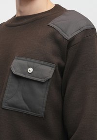 Barna pulóver bordázott textúrával, szürke steppelt vállpárnával és elülső zsebbel, amely biztonságos gombos záródással rendelkezik.
