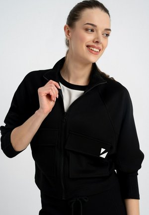 Młoda kobieta uśmiecha się, ubrana w czarną kurtkę zapinaną na zamek z dużymi kieszeniami, na białej koszuli, na tle jednolitego jasnego tła.