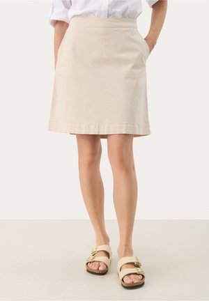 Kobieta w beżowej spódnicy do kolan z kieszeniami, białej koszuli włożonej do środka oraz beżowych sandałach na klamrę, stojąca na jednolitym tle.