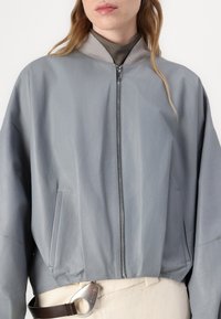 Jaqueta bomber de couro cinza com colarinho canelado, fecho em zip e bolsos laterais, combinada com um detalhe na cintura em bege.