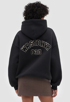 Person with curly hair wears black hoodie with "WRSTBHVR 030" text on back and a brown textured mini skirt against a plain background.