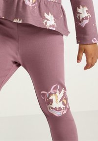 Fioletowe legginsy z miękkiego materiału, z grafiką jednorożca na prawym kolanie, ozdobione kokardką i gwiazdkami.