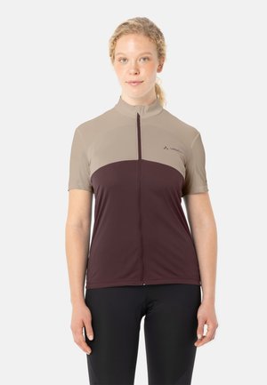 Vrouw met licht krullend haar, gekleed in een beige en donkerviolette fietstrui met korte mouwen en zwarte legging, kijkend naar voren.