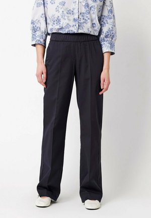 Persona che indossa pantaloni neri larghi, scarpe bianche e una camicia blu a fiori con maniche lunghe, in piedi davanti a uno sfondo bianco uniforme.