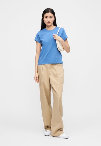 Kék rövid ujjú pamut póló, magas derekú bézs nadrággal párosítva. A modell egy kis fehér kézitáskát tart, és fehér sportcipőt visel.