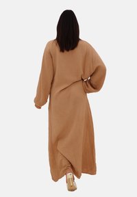 Kvinde med langt hår, der går væk iført en løstsiddende beige kjole og sandaler med stropper og hæl. Minimalistisk baggrund.