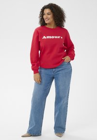 Rotes Sweatshirt mit weißem "Amour"-Schriftzug, rundem Halsausschnitt und langen Ärmeln. Kombiniert mit hoch taillierter blauer Jeans und hellen Schuhen.
