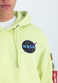 Svetlozelená mikina s kapucňou, ktorá má nášivku loga NASA, šnúrku na kapucni a zipsovú kapsu s červeným označením. Mäkká textúra látky.