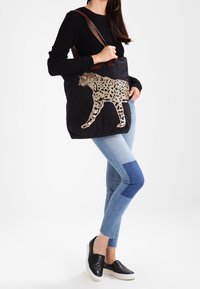 Sac fourre-tout en tissu noir avec un motif léopard beige, des poignées marron et un intérieur spacieux. La texture semble lisse avec une finition douce.