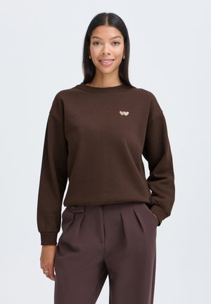 Femme aux longs cheveux noirs portant un sweat-shirt marron et un pantalon marron assorti, souriante avec une main dans la poche.