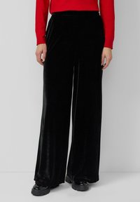 Zwarte fluwelen wijde pijpenbroek met een gladde textuur, voorzien van een hoge tailleband. Combinatie met een rood shirt met lange mouwen en chunky zwarte schoeisel.