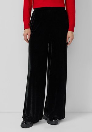 Czarne welurowe spodnie o szerokim kroju, o gładkiej teksturze, z wysokim stanem. Połączone z czerwonym topem z długim rękawem i masywnym czarnym obuwiem.