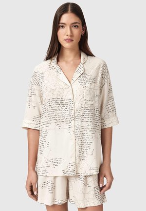 Jonge vrouw met lang haar draagt een crèmekleurig, kort mouwtjes overhemd met knopen en bijpassende korte broek, beide met zwarte schrijf- en kaartlijnpatronen.