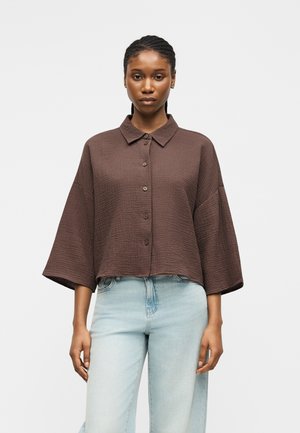 Kobieta z warkoczami, ubrana w luźną brązową koszulę na guziki z szerokimi rękawami oraz jasnoniebieskie, wysokie spodnie dżinsowe, stojąca na jednolitym tle.