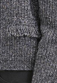 Gray knitted sweater with a ribbed texture, featuring a fringe detail on the sleeve. The fabric blends dark and light hues.