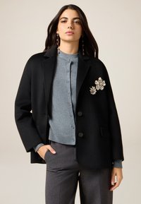 Schwarzer Wollblazer mit aufgeklapptem Revers, kombiniert mit einer hellblauen Kaschmirstrickjacke mit Knöpfen und gerippten Bündchen. Florale Verzierungen am Blazer.