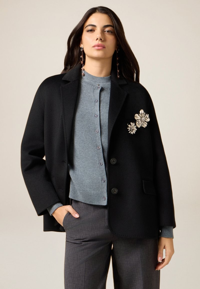 Schwarzer Wollblazer mit aufgeklapptem Revers, kombiniert mit einer hellblauen Kaschmirstrickjacke mit Knöpfen und gerippten Bündchen. Florale Verzierungen am Blazer.