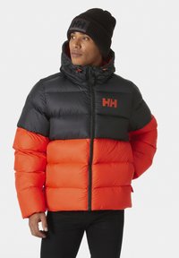 Giubbotto piumino con design trapuntato, caratterizzato dalla parte superiore nera e dalla parte inferiore arancione brillante, chiusura con zip e cappuccio. Accompagnato da un berretto in maglia nero.