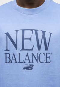 Άτομο που φοράει ανοιχτό μπλε φούτερ με μεγάλο σκούρο μπλε κείμενο "NEW BALANCE" και λογότυπο στο κέντρο του στήθους.