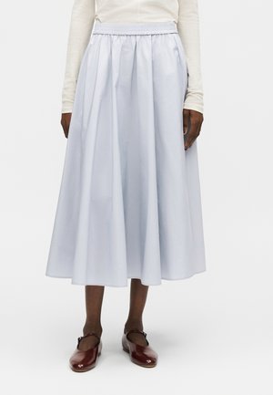 Naine, kes kannab valget pika varrukaga toppi, helehalli kogutud midi seelikut elastse vöökohaga ja tumepunaseid Mary Jane tüüpi kingi, seisab.