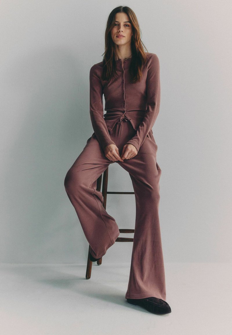 Junge Frau mit langen braunen Haaren, die ein altrosa geripptes, geknöpftes Oberteil und passende weite Hosen trägt, sitzt auf einem Holzstuhl vor schlichtem Hintergrund.