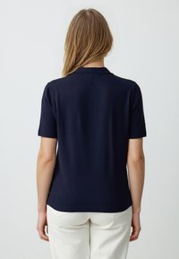 Granatowa, krótkozasięgowa koszulka polo z prążkowanym kołnierzykiem i wykończeniem, wykonana z gładkiego materiału, noszona z jasnymi spodniami.