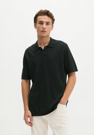 Jeune homme aux cheveux ondulés portant un polo vert foncé à manches courtes et un pantalon beige clair, debout devant un fond blanc uni.