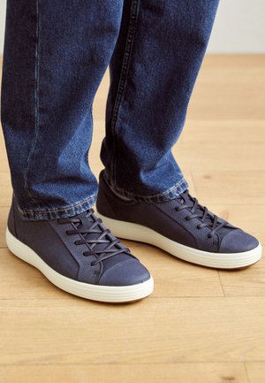 Dunkelblaue Sportschuhe mit strukturiertem Material, weißen Sohlen und flachen Schnürsenkeln, kombiniert mit dunkelblauen Jeans auf einem Holzboden.