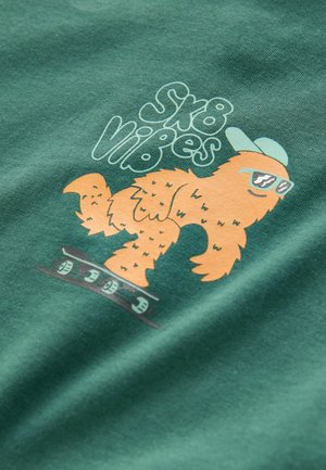 Zöld póló, rajzfilmfigúrával, narancssárga szőrzettel, napszemüveget és sapkát viselve, valamint a „Sk8 Vibes” szöveg játékos betűtípussal.