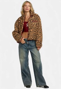 Giacca corta arancione e nera con motivo a fantasia, colletto alto e tasca con cerniera, abbinata a jeans blu larghi. Tessuto morbido e strutturato.