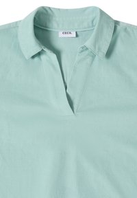 Chemise vert clair à manches courtes avec un col et une ouverture en V, confectionnée en tissu doux, étiquetée "CECIL" à l'intérieur du col.