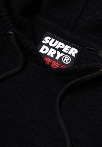 Felpa nera con tessuto testurizzato; presenta un'etichetta logo prominente con "SUPER DRY" in bianco e rosso. Include un cappuccio con coulisse.