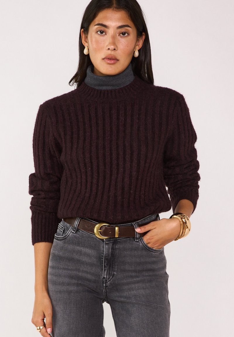 Donna con maglione a coste scuro sopra un dolcevita grigio, jeans grigi con cintura marrone, braccialetto d'oro, anello e orecchini a goccia.