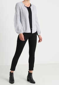 Mujer vistiendo un cárdigan abierto de manga larga gris claro, top negro, pantalones ajustados negros y botines negros, de pie contra un fondo plano.