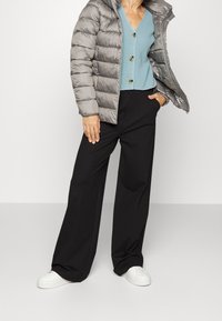 Graue Steppjacke mit Kapuze, hellblaues Strickoberteil mit Knöpfen, schwarze Hosen mit weitem Bein und weiße Sneaker.