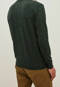 Tmavě zelený pletený svetr s texturovaným vzorem, dlouhými rukávy a zaobleným lemem, kombinovaný s hnědými kalhotami.