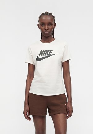 Jeune femme aux cheveux tressés portant un t-shirt blanc Nike et un short marron, debout devant un fond uni clair.