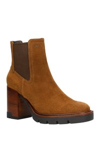 Stivaletto alla caviglia in suede marrone con punta arrotondata, pannelli laterali elastici e tacco in legno impilato. Presenta cuciture a contrasto e suola in gomma.