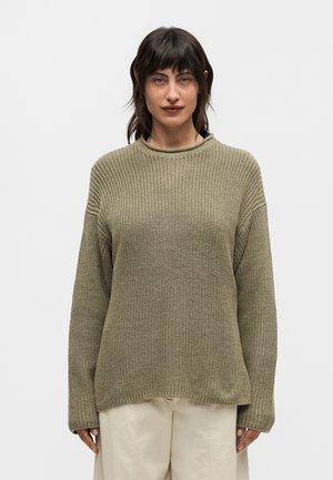 Γυναίκα με σκούρα μαλλιά που φοράει ένα φαρδύ πλεκτό πουλόβερ σε ελιά πράσινο χρώμα και μπεζ παντελόνι, στέκεται μπροστά σε ένα απλό φωτεινό φόντο.