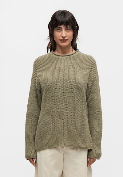 Femme aux cheveux foncés portant un pull en maille vert olive oversize et un pantalon beige, debout devant un fond clair uni.
