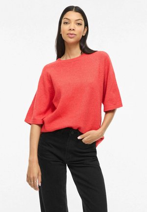 Nő áll zsebében tartott kézzel, laza piros rövid ujjú pulóvert és fekete magasderekú nadrágot viselve fehér háttér előtt.