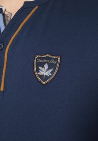 Navy blue polo shirt featuring a short placket, brown accents, and an embroidered shield logo with "Charles Colby" and a leaf design.