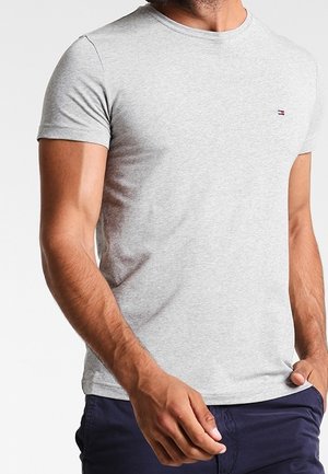 Mann trägt eng anliegendes hellgraues T-Shirt mit kurzen Ärmeln und kleinem Logo auf der Brust sowie dunkelblaue Hose, Hand in der Nähe der Tasche.